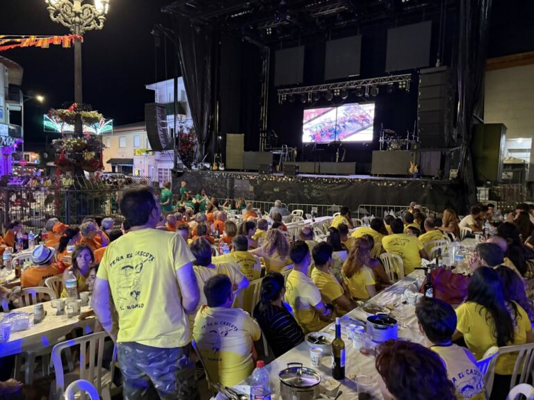 Multitud de personas sentadas en mesas al aire libre por la noche, disfrutando de una cena popular frente a un gran escenario iluminado durante las Fiestas de El Boalo.