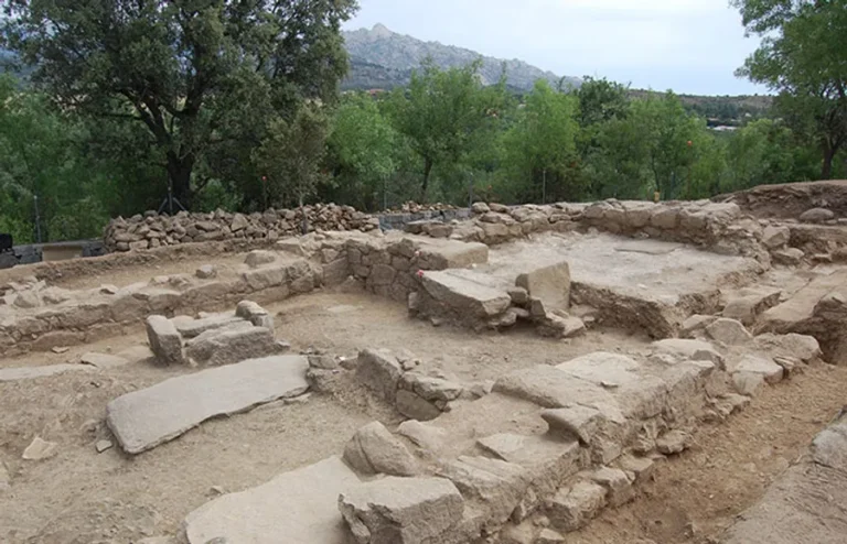 Vestigios de un yacimiento arqueológico de piedra con muros y tumbas, en un entorno de tierra y vegetación, con las montañas de la sierra al fondo.
