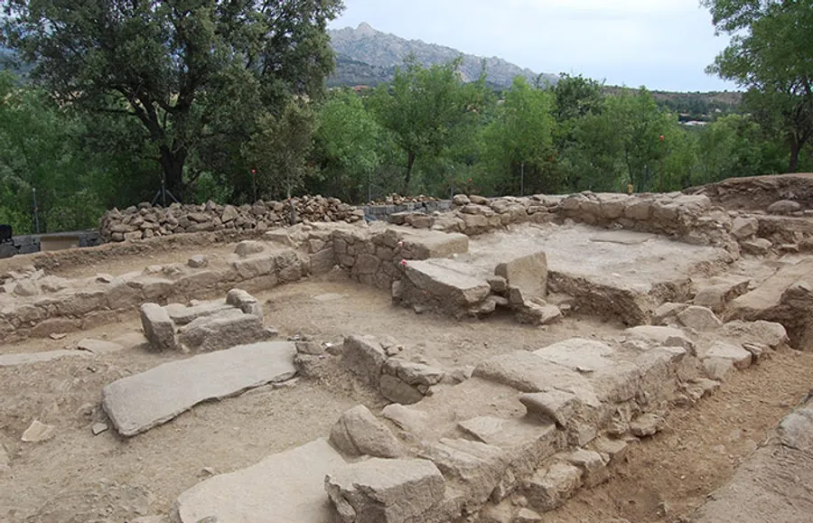 Vestigios de un yacimiento arqueológico de piedra con muros y tumbas, en un entorno de tierra y vegetación, con las montañas de la sierra al fondo.
