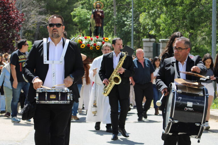 Miembros de una banda de música, tocando tambores y saxofón, lideran la procesión de San Isidro en El Boalo, con la estatua del santo visible al fondo.