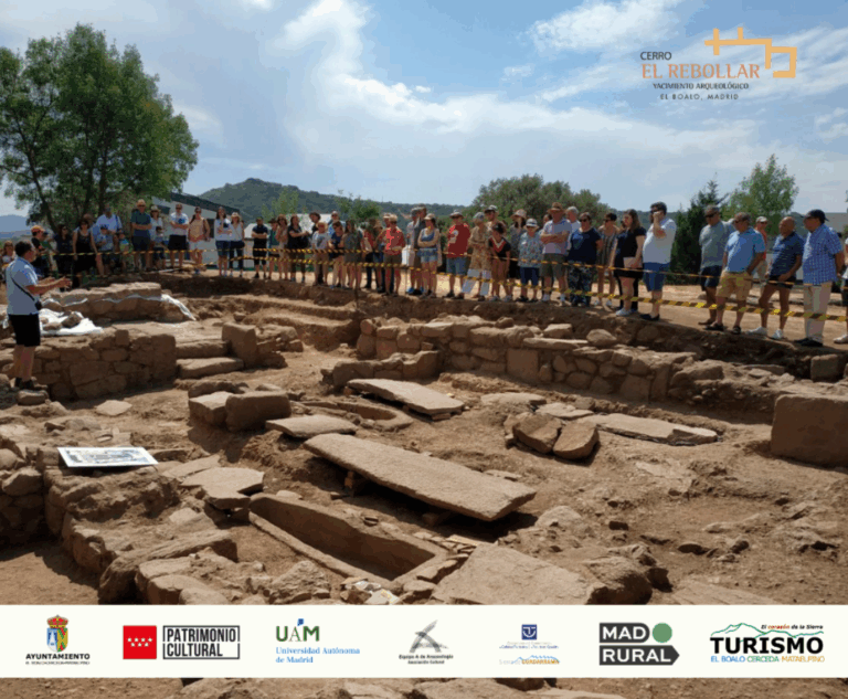 Un grupo de personas en una visita guiada a un yacimiento arqueológico al aire libre, con una persona explicando y estructuras de piedra y tumbas.