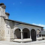 Fachada de piedra de la Iglesia de San Sebastián Mártir en El Boalo, con campanario y nidos de cigüeñas, bajo un cielo azul.