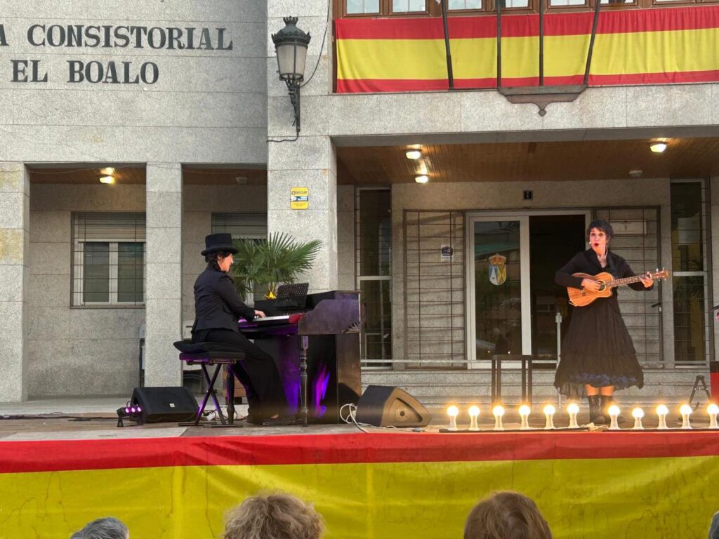 Dos artistas actúan en un escenario al aire libre frente a un edificio con banderas españolas, una tocando el piano y otra cantando y tocando un ukelele.