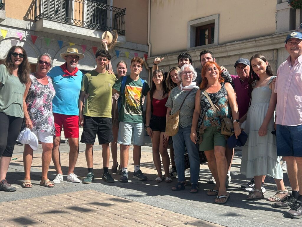 Un grupo diverso de personas, incluyendo locales y posibles extras, posan sonrientes en una calle decorada con banderines, durante el rodaje de una película.