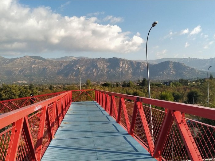 Pasarela peatonal de metal pintada de rojo y azul con vista a las montañas de la Sierra de Guadarrama bajo un cielo azul con nubes blancas.