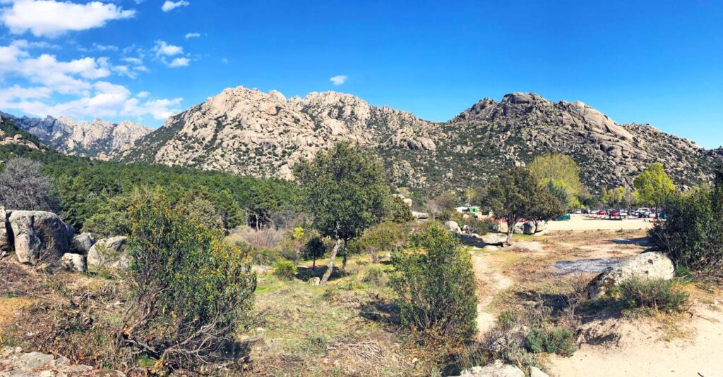Amplia vista panorámica de La Pedriza con formaciones rocosas graníticas, vegetación mediterránea y un área de estacionamiento al fondo bajo un cielo azul.