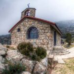 Vista frontal de la Ermita de San Isidro Labrador en El Boalo, una pequeña capilla de piedra con techo de tejas, campanario y una ventana con rejas, en un día nublado.