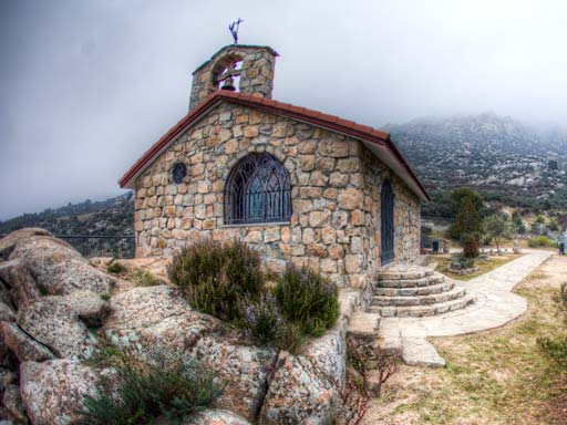 Vista frontal de la Ermita de San Isidro Labrador en El Boalo, una pequeña capilla de piedra con techo de tejas, campanario y una ventana con rejas, en un día nublado.