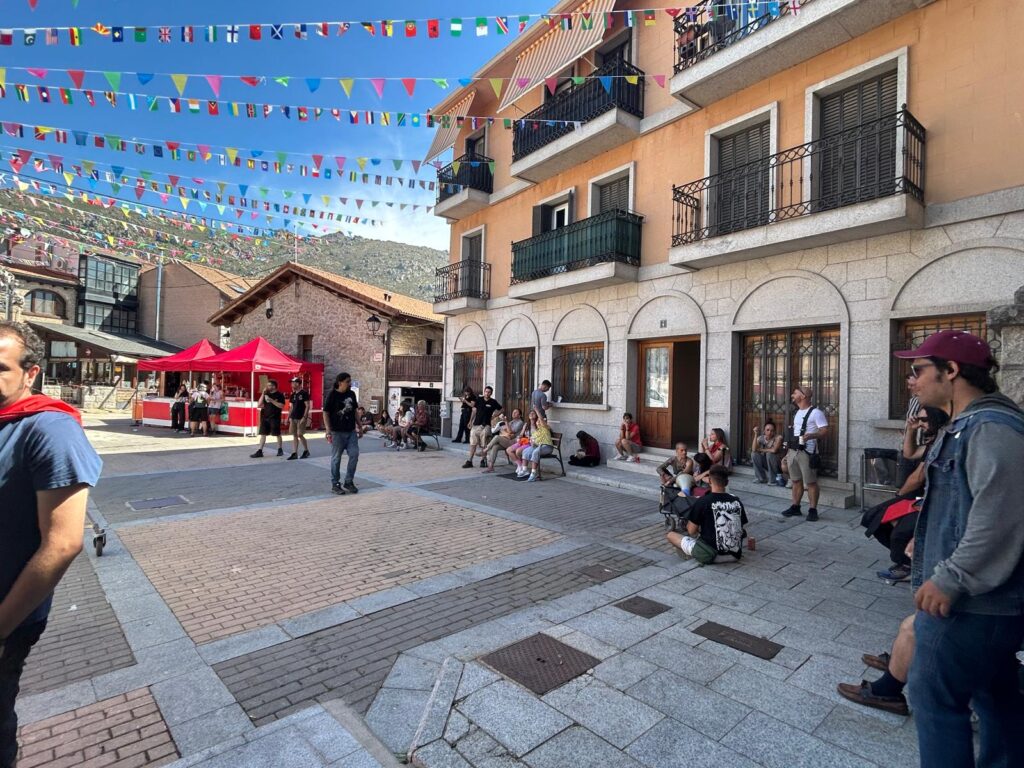 Una plaza de pueblo adornada con banderines de colores, con personas reunidas y algunas carpas rojas.