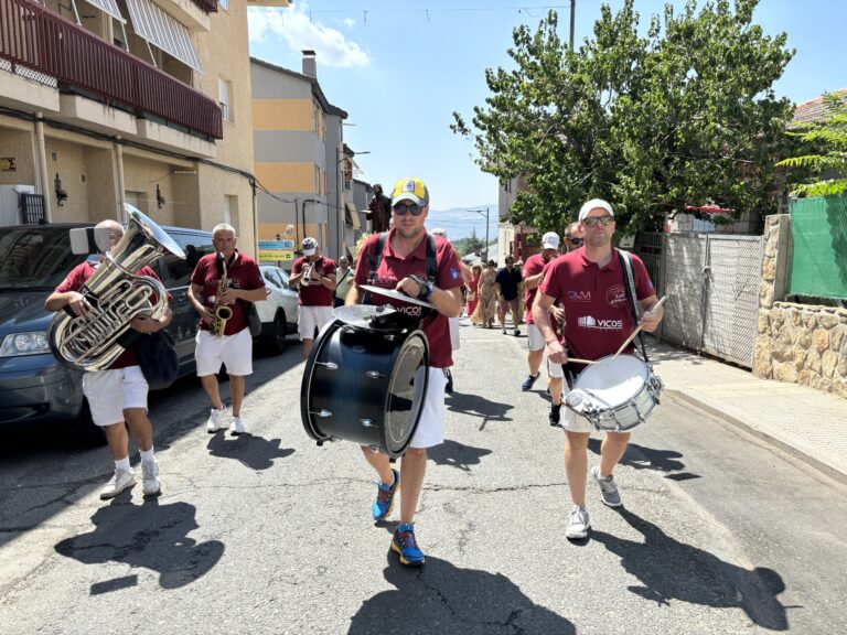 Banda musical desfilando por una calle soleada de Mataelpino durante sus Fiestas de Verano, con músicos tocando instrumentos de viento y percusión.