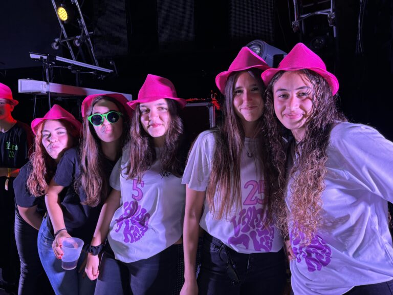Grupo de jóvenes, algunas con sombreros fucsias y camisetas de fiesta, posando sonrientes en un evento nocturno con iluminación de escenario, durante las Fiestas de Verano de Cerceda.