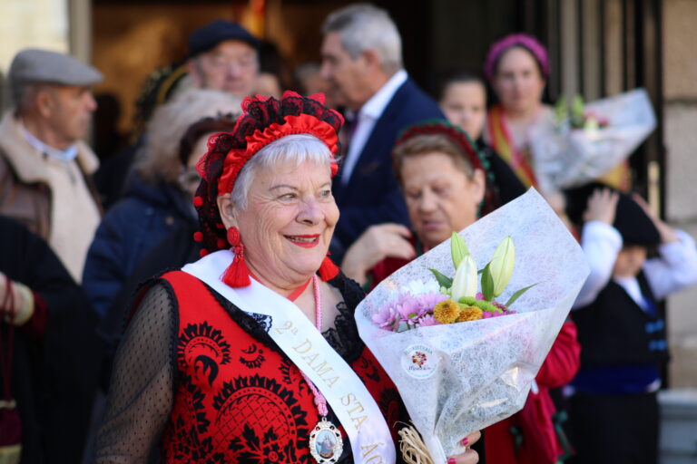 Una mujer sonriente, de edad avanzada, vestida con traje tradicional y una banda con la inscripción "2ª Dama Sta. Águeda", sosteniendo un ramo de flores.