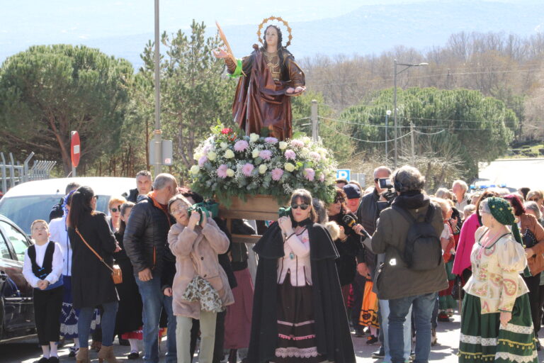 La estatua de Santa Águeda es transportada en procesión por una multitud de personas, algunas con trajes tradicionales, por las calles de Mataelpino, con montañas al fondo.