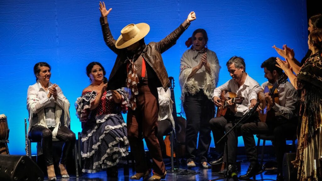 Fotografía de una actuación de flamenco en un escenario con un bailarín en el centro, rodeado de músicos y otros artistas.