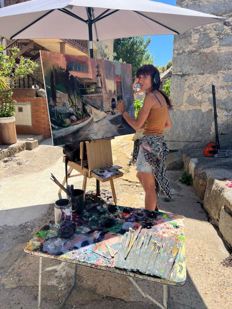 Una joven artista con auriculares pintando un paisaje urbano en un lienzo sobre un caballete, en una calle empedrada bajo un paraguas de sol.