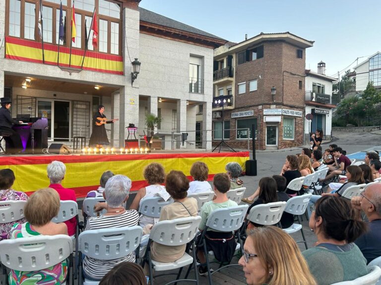 Vista trasera de una audiencia sentada en sillas blancas, observando un escenario al aire libre donde actúan dos artistas frente a un edificio con banderas.