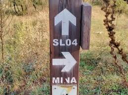 Poste de madera con señalización de ruta de senderismo, mostrando flechas blancas con códigos "SL04" y "MINA" en un entorno de vegetación seca.