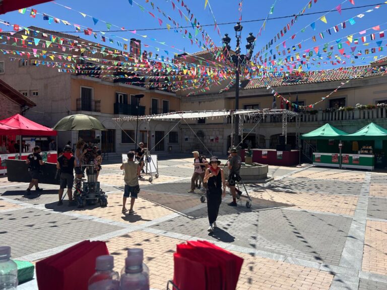 Una plaza de pueblo con banderines de colores, equipo de rodaje y carpas, bajo un cielo azul brillante.