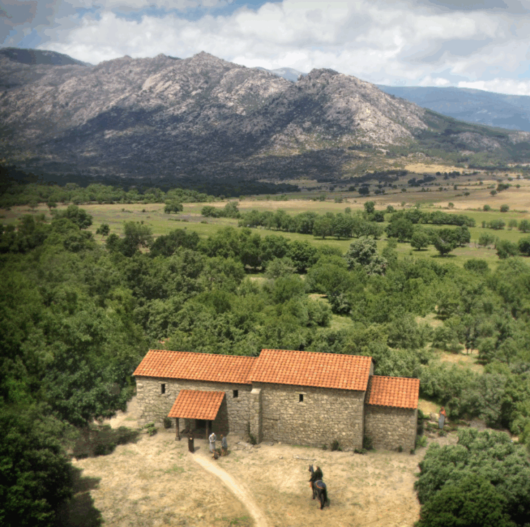 Reconstrucción aérea de una iglesia visigoda de piedra y tejas rojas, rodeada de un paisaje rural con árboles y un valle, con las montañas de la Sierra de Guadarrama al fondo.