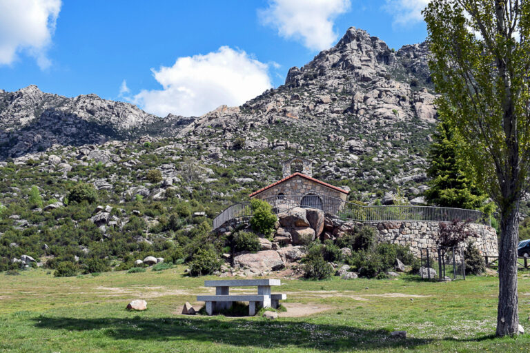 Ermita de San Isidro Labrador en El Boalo en un entorno natural amplio, con bancos de piedra, montañas rocosas al fondo y cielo azul con nubes.