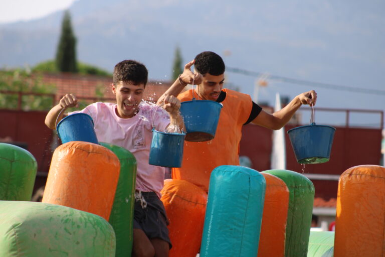 Dos jóvenes sonriendo mientras salpican agua con cubos azules, de pie entre obstáculos hinchables de colores, durante una actividad acuática en las Fiestas de Verano de Cerceda.