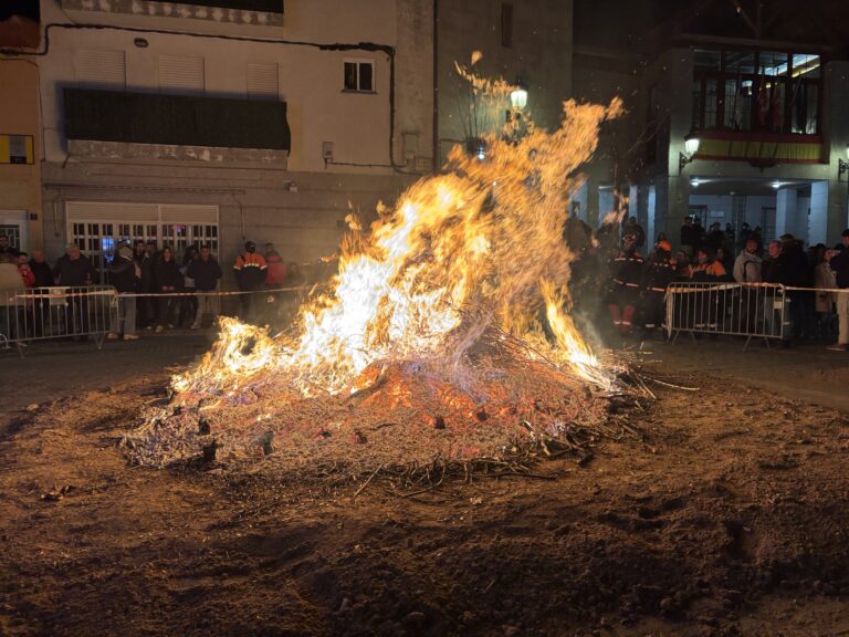Una gran hoguera ardiendo intensamente en la calle por la noche, con espectadores y personal de seguridad detrás de vallas, durante la celebración de "La Luminaria" en El Boalo, Cerceda y Mataelpino.
