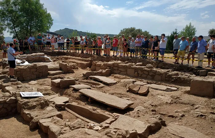 Grupo de personas observando un yacimiento arqueológico guiadas por un experto, con muros de piedra y tumbas a la vista.