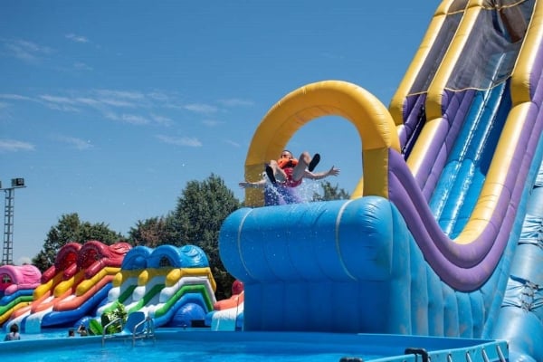 Una persona con chaleco salvavidas naranja deslizándose por un gran tobogán hinchable azul y amarillo hacia una piscina de AguaPark Toledo, con el agua salpicando.