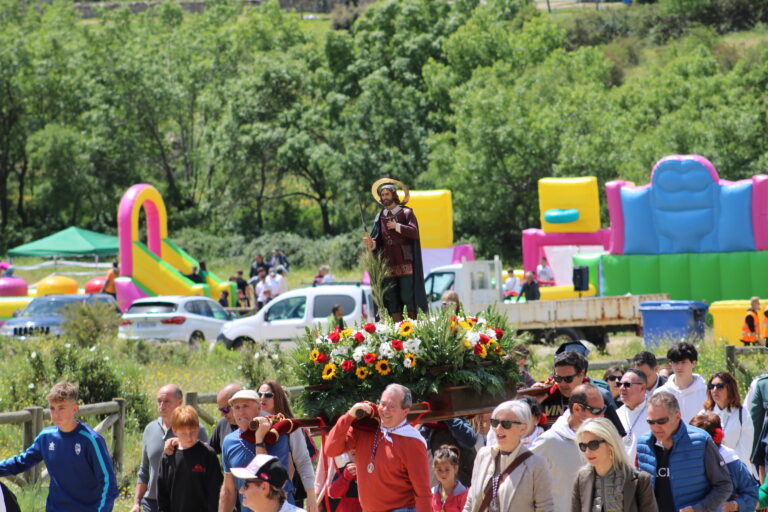 La estatua de San Isidro Labrador es portada en procesión por un grupo de hombres en un entorno campestre, con castillos hinchables de colores visibles al fondo, en la Romería de El Boalo.