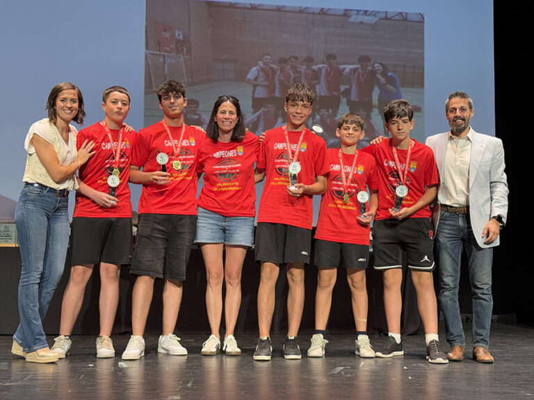 Grupo de jóvenes atletas masculinos, vestidos con camisetas rojas de "CAMPEONES", mostrando sus medallas y trofeos en un escenario, con dos entrenadoras o colaboradoras.