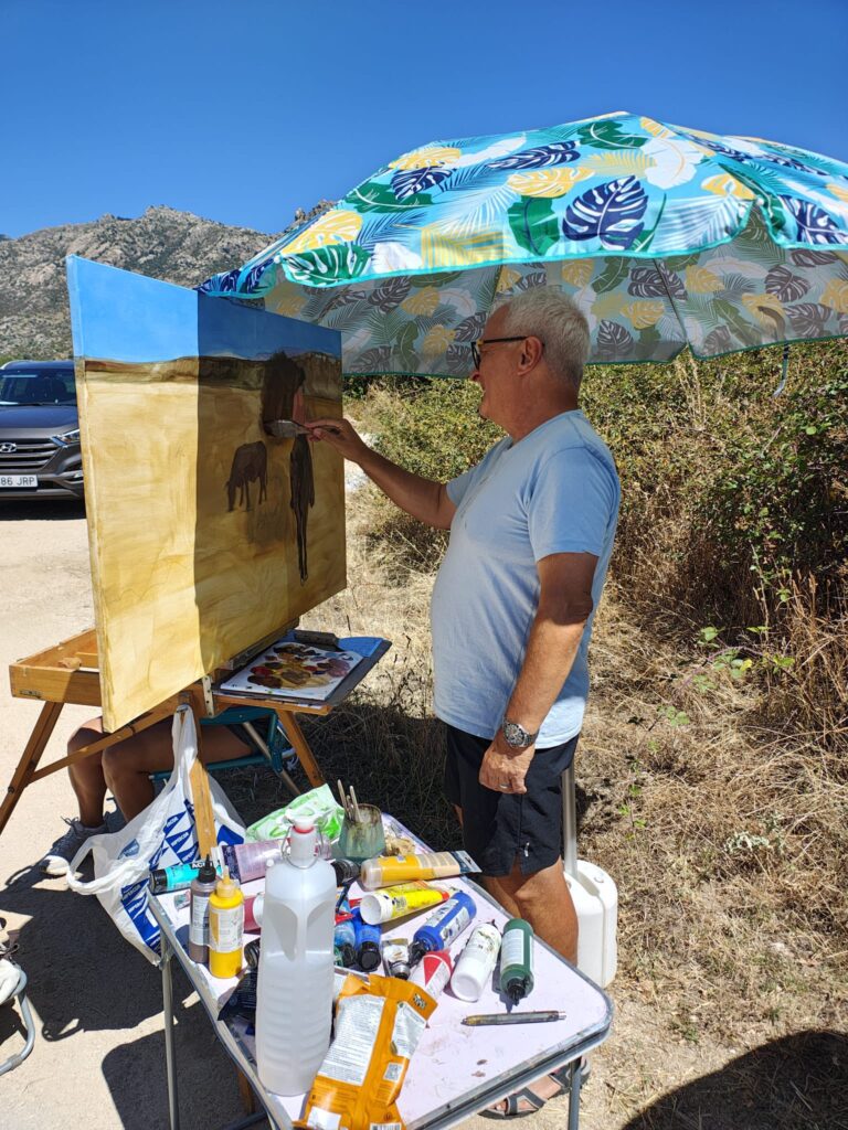 Un hombre mayor con gafas de sol y gorra pintando un paisaje rural con árboles y montañas en un lienzo sobre un caballete, bajo una sombrilla colorida.