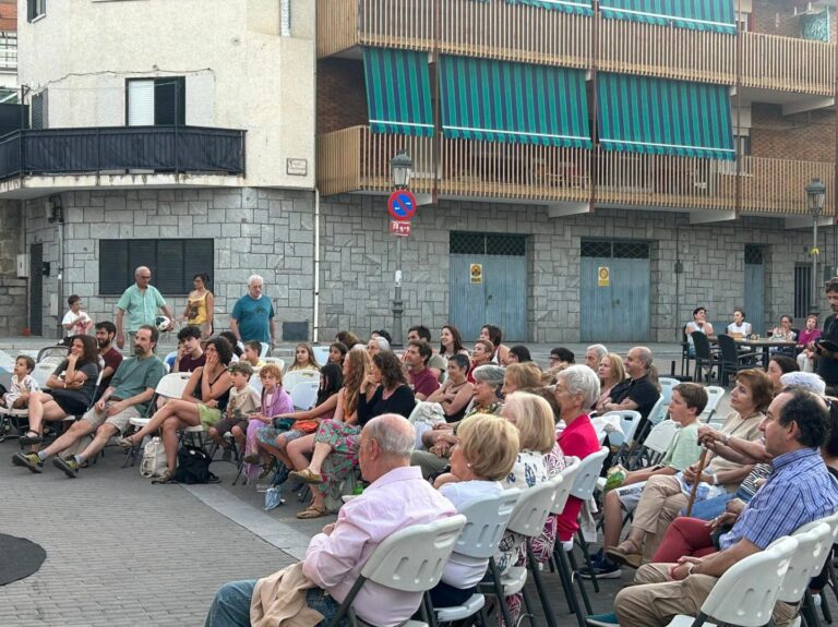 Grupo diverso de personas, jóvenes y mayores, sentadas en sillas blancas en una plaza, observando una actuación artística al aire libre durante el atardecer.