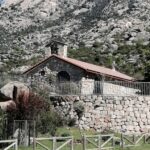Ermita de San Isidro Labrador en El Boalo vista desde abajo, con la fachada de piedra, el tejado, una valla, y las imponentes montañas rocosas de La Pedriza al fondo.