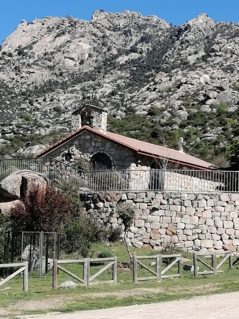 Ermita de San Isidro Labrador en El Boalo vista desde abajo, con la fachada de piedra, el tejado, una valla, y las imponentes montañas rocosas de La Pedriza al fondo.