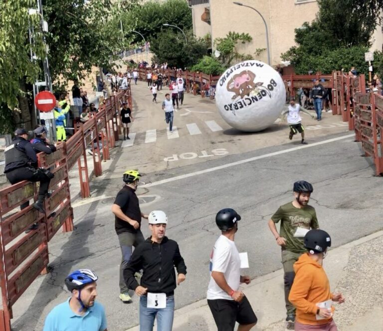 Varias personas, algunas con cascos, corriendo por una calle empinada mientras una gran bola blanca del "Boloencierro Mataelpino" rueda detrás de ellas.