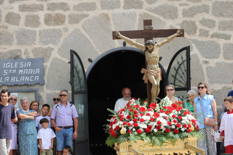 Una procesión con la estatua de Cristo en la cruz, adornada con flores, saliendo de la Iglesia Santa María La Blanca en Cerceda, un acto de fe y tradición en las Fiestas de Verano.