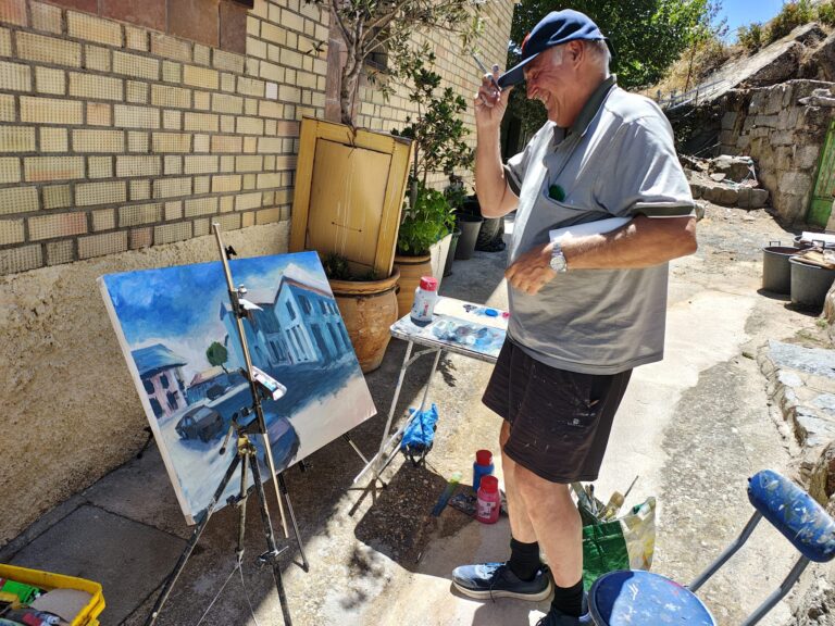 Un hombre mayor con gorra, sonriendo y sosteniendo un cuadro de paisaje urbano con tonos azules, con un caballete y material de pintura a su lado.
