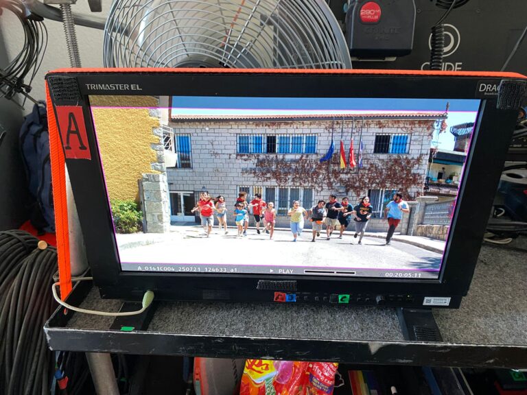 Un monitor de cine muestra una toma de una película en progreso, con un grupo de personas corriendo en una calle de un pueblo, con banderas en las casas.