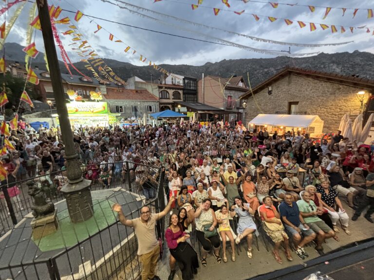 Una plaza de Mataelpino abarrotada de gente de noche, con guirnaldas de luces, un escenario al fondo y montañas a la distancia, durante las Fiestas de Verano.