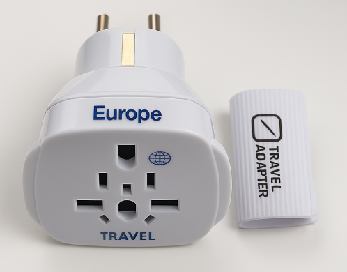 Un adaptador de viaje universal blanco, con inscripciones "Europe" y "Travel", y una pieza accesoria con el texto "TRAVEL ADAPTER", sobre una superficie clara.
