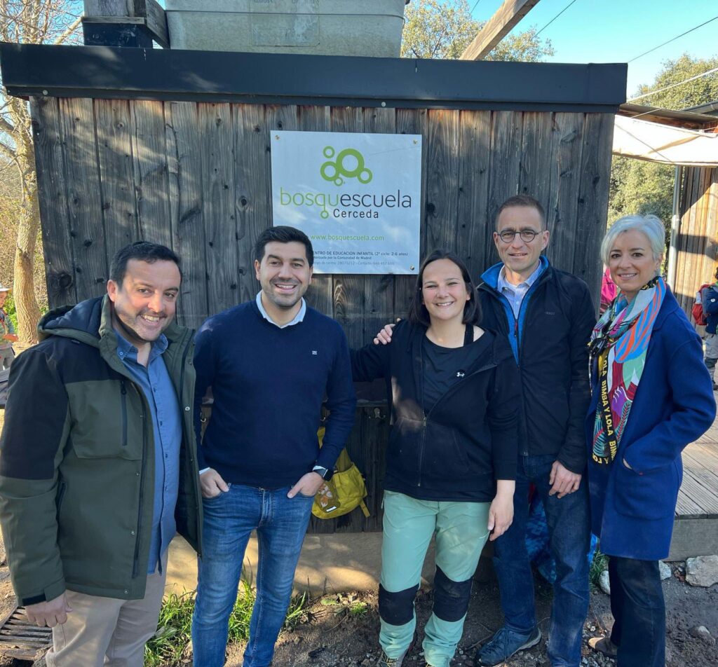 Grupo de adultos, incluyendo al alcalde y concejal de educación de El Boalo, Cerceda y Mataelpino, posando frente al cartel de Bosquescuela Cerceda, símbolo de la innovación educativa apoyada por el Ayuntamiento.