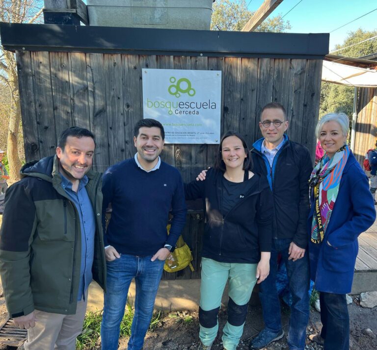 Grupo de adultos, incluyendo al alcalde y concejal de educación de El Boalo, Cerceda y Mataelpino, posando frente al cartel de Bosquescuela Cerceda, símbolo de la innovación educativa apoyada por el Ayuntamiento.