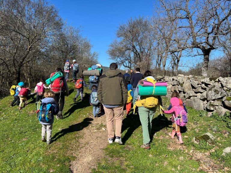 Niños y adultos con mochilas y colchonetas caminando por un sendero rural, representando las actividades al aire libre de Bosquescuela Cerceda, un proyecto valorado por el Ayuntamiento de El Boalo, Cerceda y Mataelpino.
