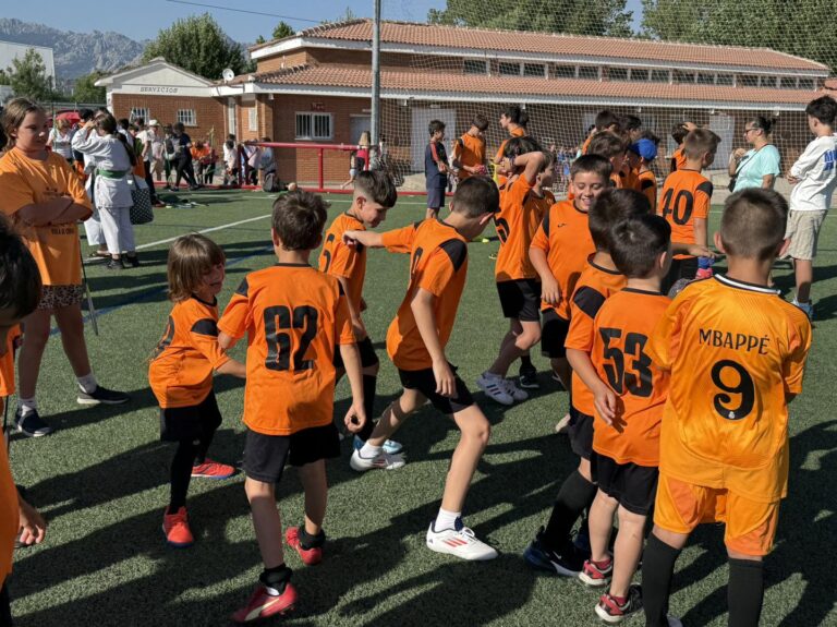 Grupo de niños con camisetas de fútbol naranjas, algunos con el nombre "Mbappé", jugando en un campo de césped artificial, simbolizando el fomento del deporte base por el Ayuntamiento de El Boalo, Cerceda y Mataelpino.