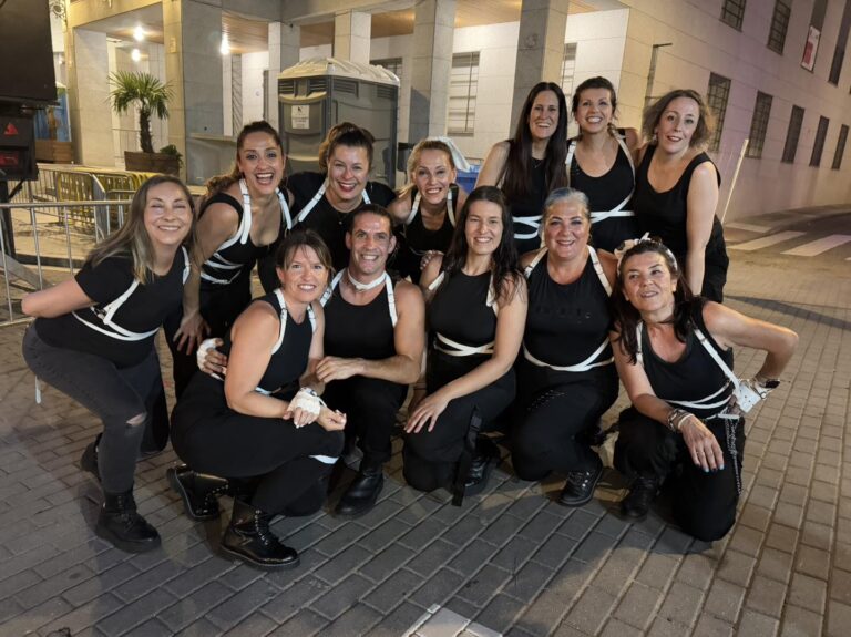 Un grupo de bailarinas y un hombre, todos con trajes negros y detalles blancos, posando sonrientes en una calle por la noche, parte de las actuaciones de las Fiestas de El Boalo 2025.