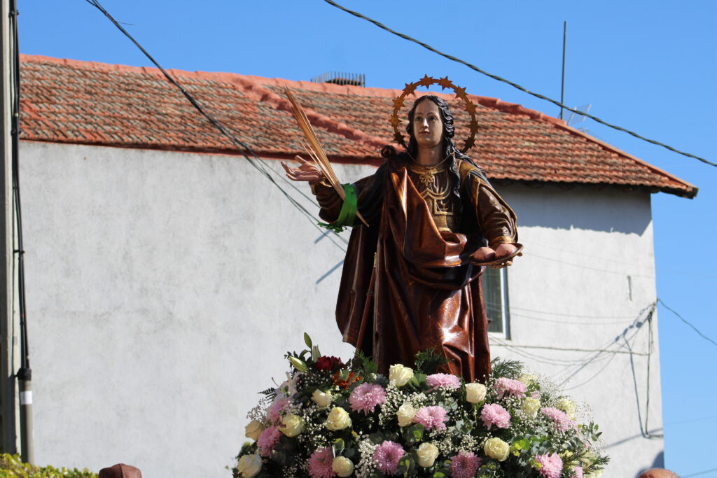 Estatua de Santa Águeda, adornada con flores y con su halo, siendo transportada durante una procesión, con una casa de fondo, en Mataelpino.