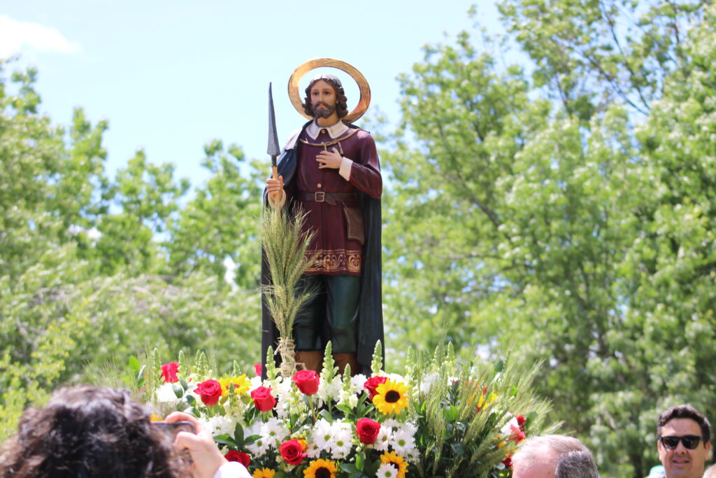 La estatua de San Isidro Labrador, con su halo y un arado, adornada con flores y espigas de trigo, es portada en procesión bajo un cielo azul, rodeada de vegetación en El Boalo.