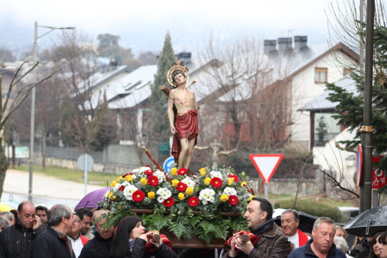 Una procesión en la calle con una estatua de San Sebastián adornada con flores, portada por hombres y mujeres, con casas y montañas cubiertas de nieve al fondo, durante las Fiestas de San Sebastián en El Boalo, Cerceda y Mataelpino.