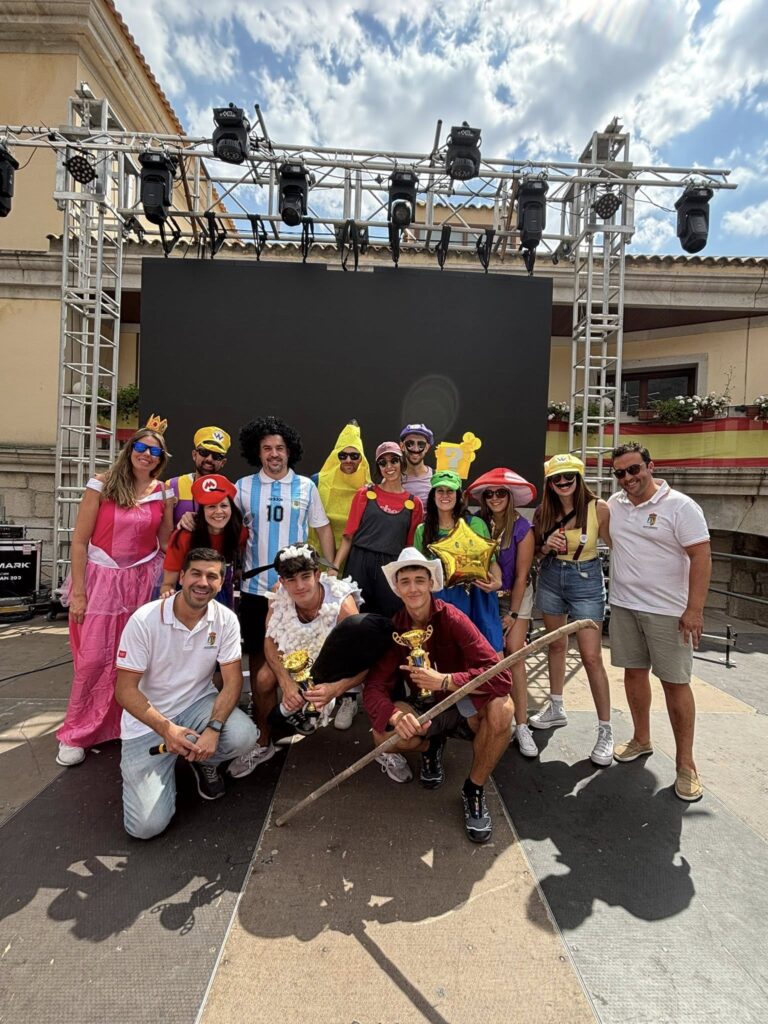 Un grupo numeroso de personas, disfrazadas de personajes de videojuegos, frutas y otros atuendos, posan sonrientes en un escenario al aire libre, con trofeos y un cetro, durante un evento festivo.