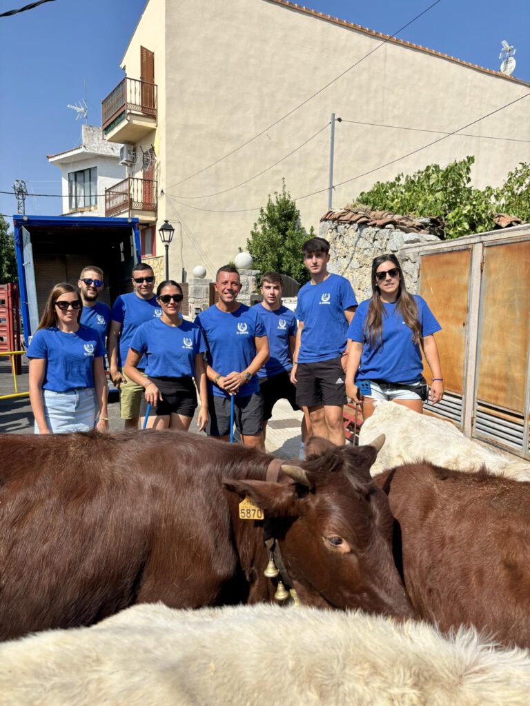 Un grupo de jóvenes, con camisetas azules con un logo, posan junto a varias vacas o toros detrás de una valla, en una calle de un pueblo, con un camión de transporte de ganado al fondo.